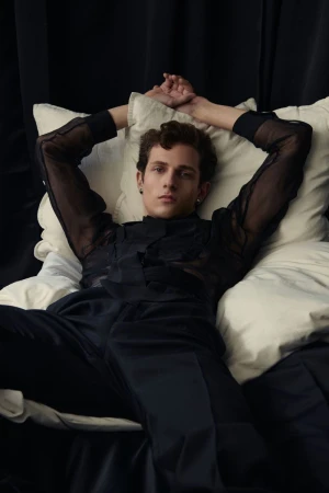 Model Mattia Liam reclining on white pillows in a black sheer shirt for Man About Town photographed by Alfonso Anton Cornelis.