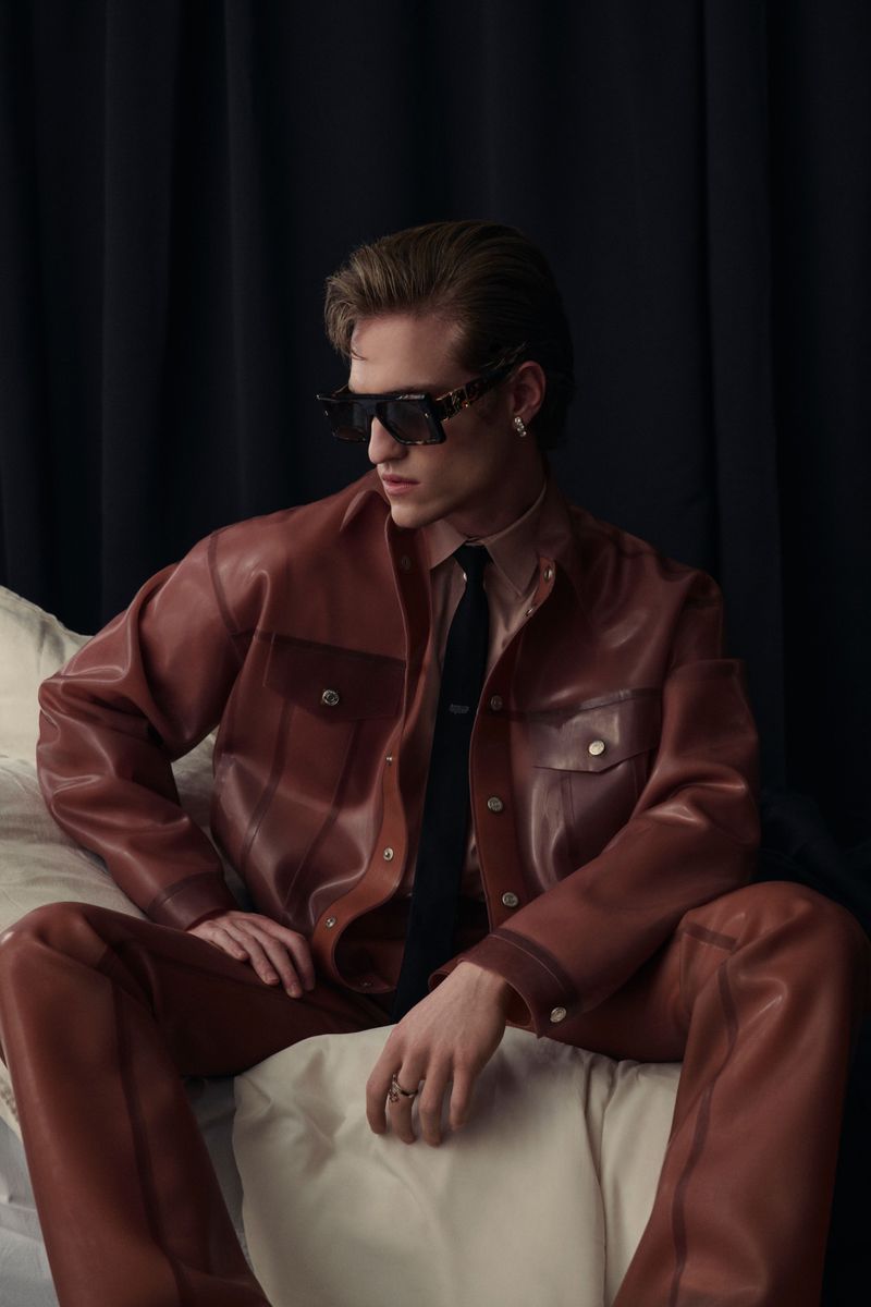 A young adult male poses indoors on a cushion, wearing a brown leather jacket, matching trousers, and sunglasses against a dark curtain backdrop.