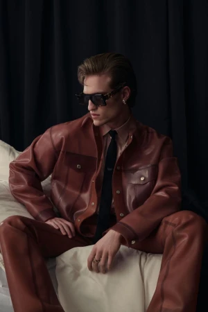 A young adult male poses indoors on a cushion, wearing a brown leather jacket, matching trousers, and sunglasses against a dark curtain backdrop.