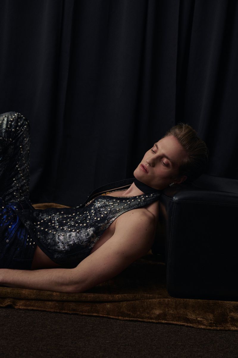 Adult male laying down in studded leather outfit with black fabric background and brown rug in eye-level indoor shot.