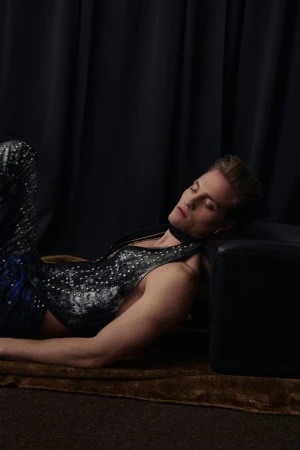 Adult male laying down in studded leather outfit with black fabric background and brown rug in eye-level indoor shot.