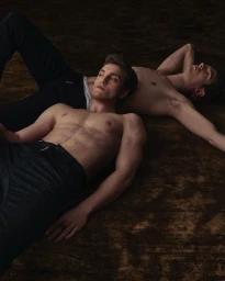 Two shirtless male models lying on a dark textured floor for Man About Town magazine