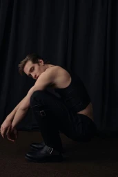 Male model crouching in a black tank top and boots against a dark curtain backdrop