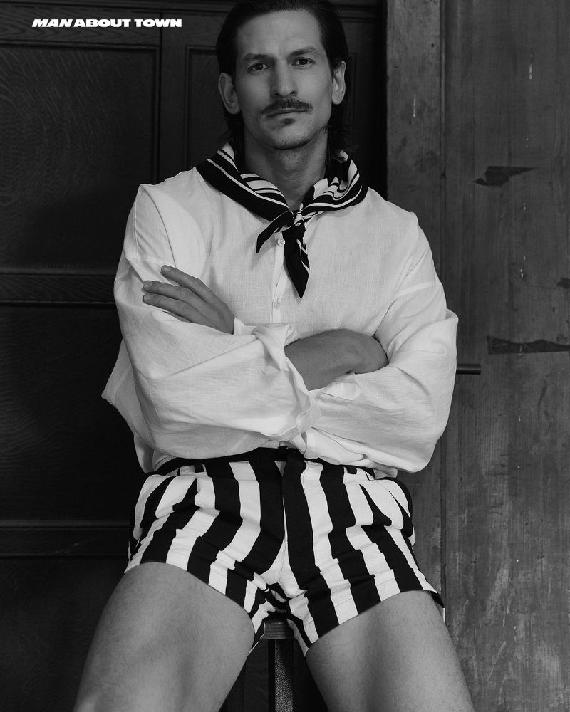 Black and white portrait of a man with a mustache wearing a sailor-style top and striped shorts standing in front of a wooden door.