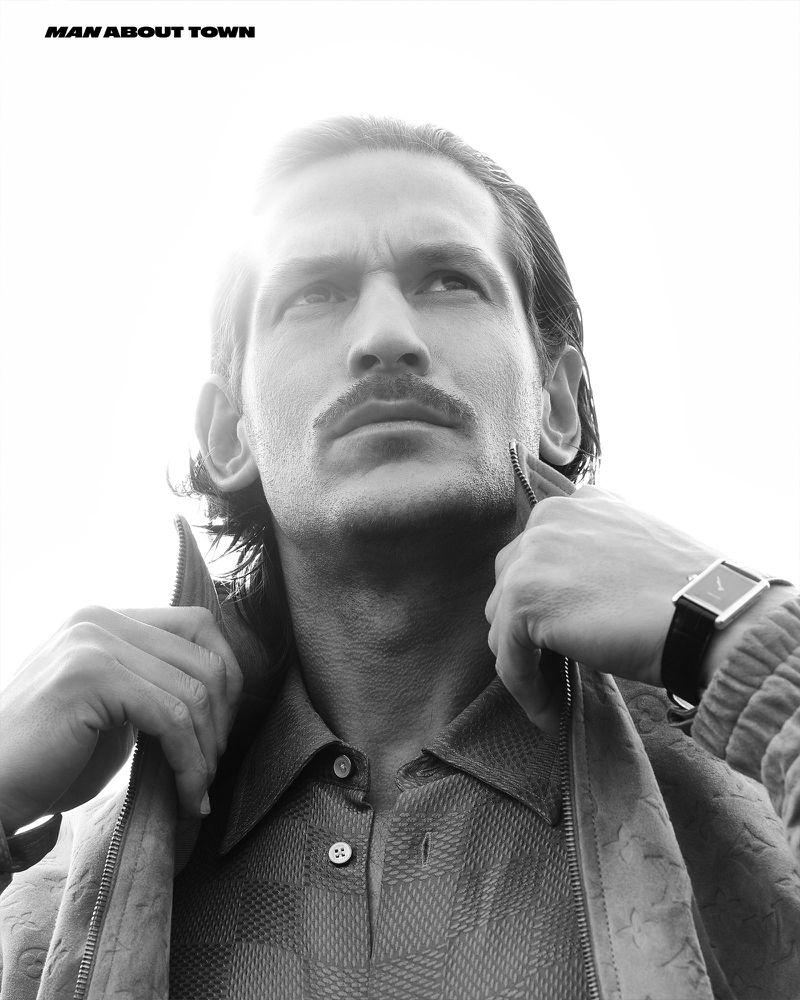 Low angle monochrome portrait of model Jarrod Scott wearing a patterned shirt and jacket with a wristwatch against a bright sky background.