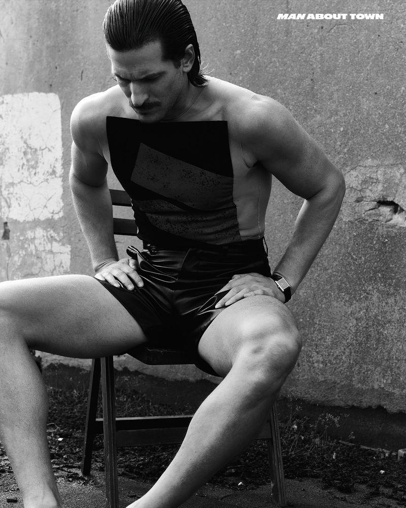 Black-and-white fashion portrait of a muscular adult male with a mustache, seated on a chair, wearing a vest and shorts.