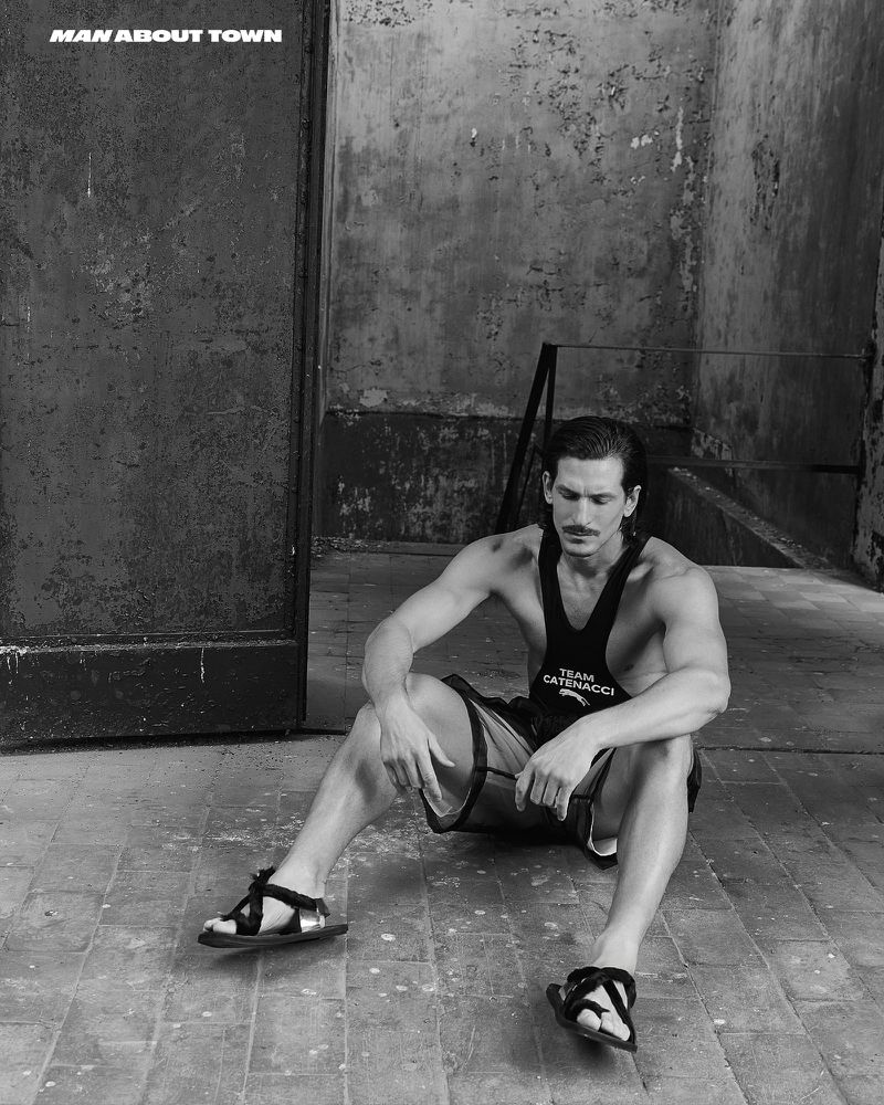 A man seated on stone tiles wearing a dark tank top and sandals against a weathered industrial wall.