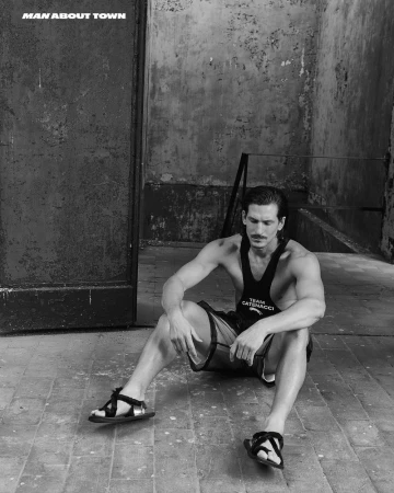A man seated on stone tiles wearing a dark tank top and sandals against a weathered industrial wall.