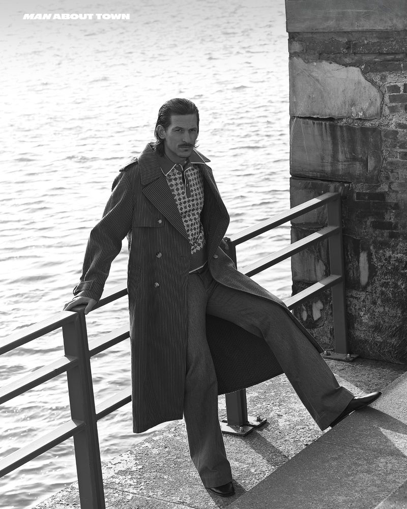 A male model poses outdoors in a long coat, dress pants and dress shoes, leaning on a metal railing with a brick wall behind him.