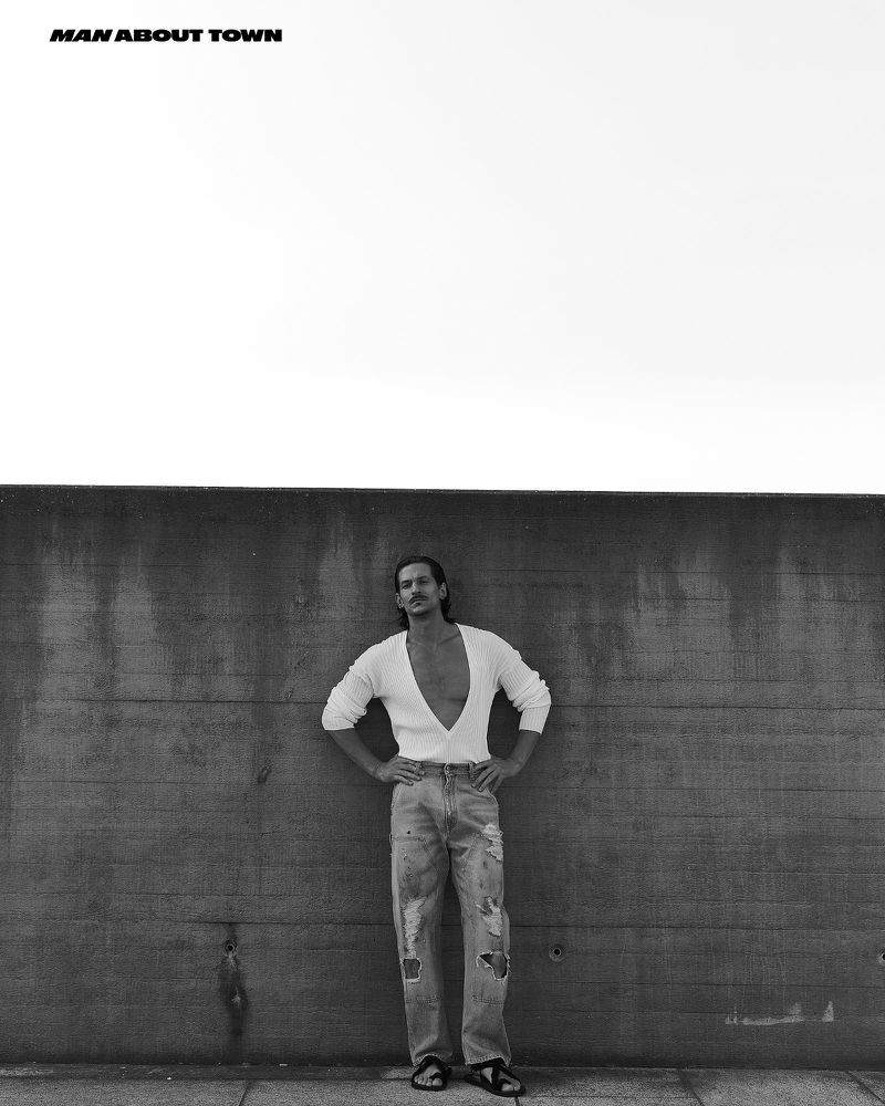 Jarrod Scott wearing a white V-neck sweater and distressed jeans standing against a dark concrete wall for Man About Town.