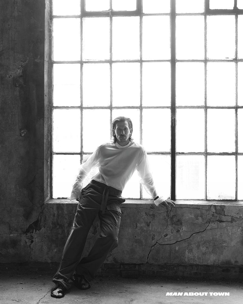 Monochrome portrait of a man leaning against a large window in a grunge, industrial-style setting, creating a high-contrast, editorial image.