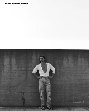 Jarrod Scott wearing a white V-neck sweater and distressed jeans standing against a dark concrete wall for Man About Town.