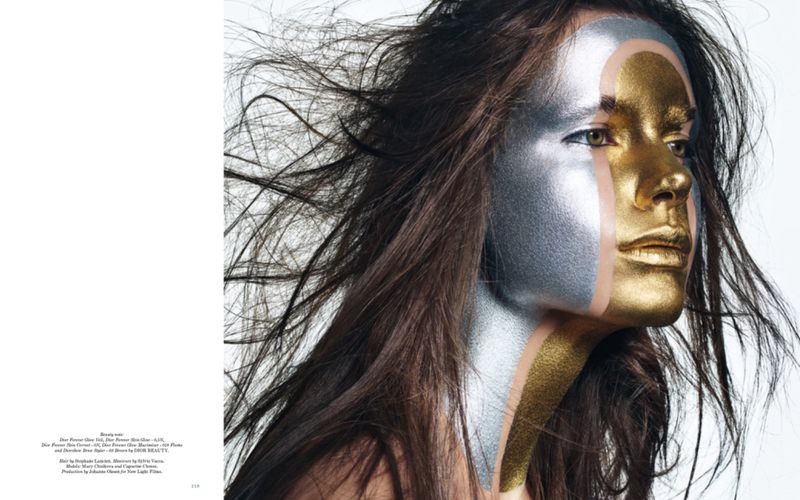 Striking beauty shot of a young adult female model with gold and silver makeup, brown hair blowing, against a bright white backdrop.