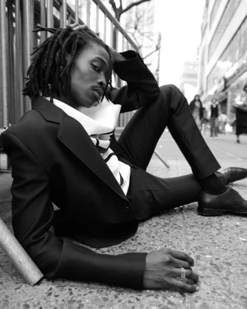Black man with dreadlocks wearing a dark suit jacket and trousers sitting on a city sidewalk leaning against a metal railing