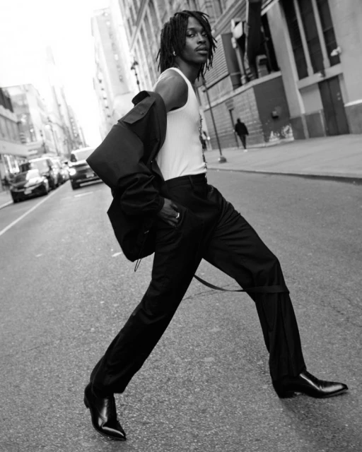 Black and white portrait of an adult man walking on a city street wearing a white tank top and black pants with cars and buildings in the background.