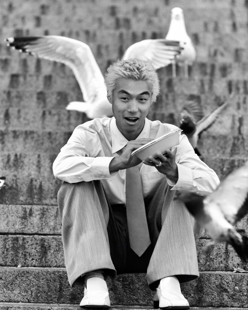Monochrome portrait of an Asian man seated on stone steps surrounded by flying seagulls while holding a dish of food for the birds.