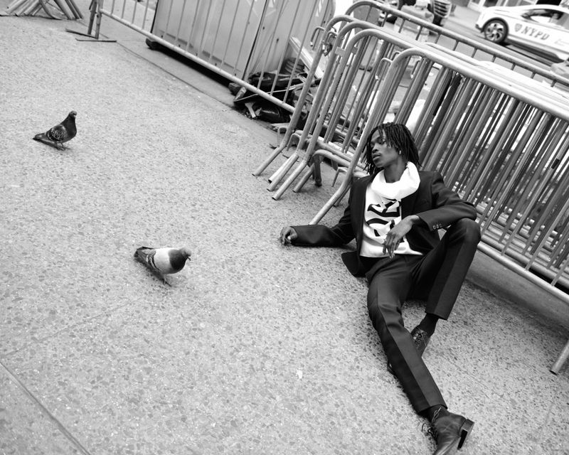 Man lies on concrete by metal barriers near two pigeons, wearing a dark suit, white shirt, and dark shoes in an urban setting.