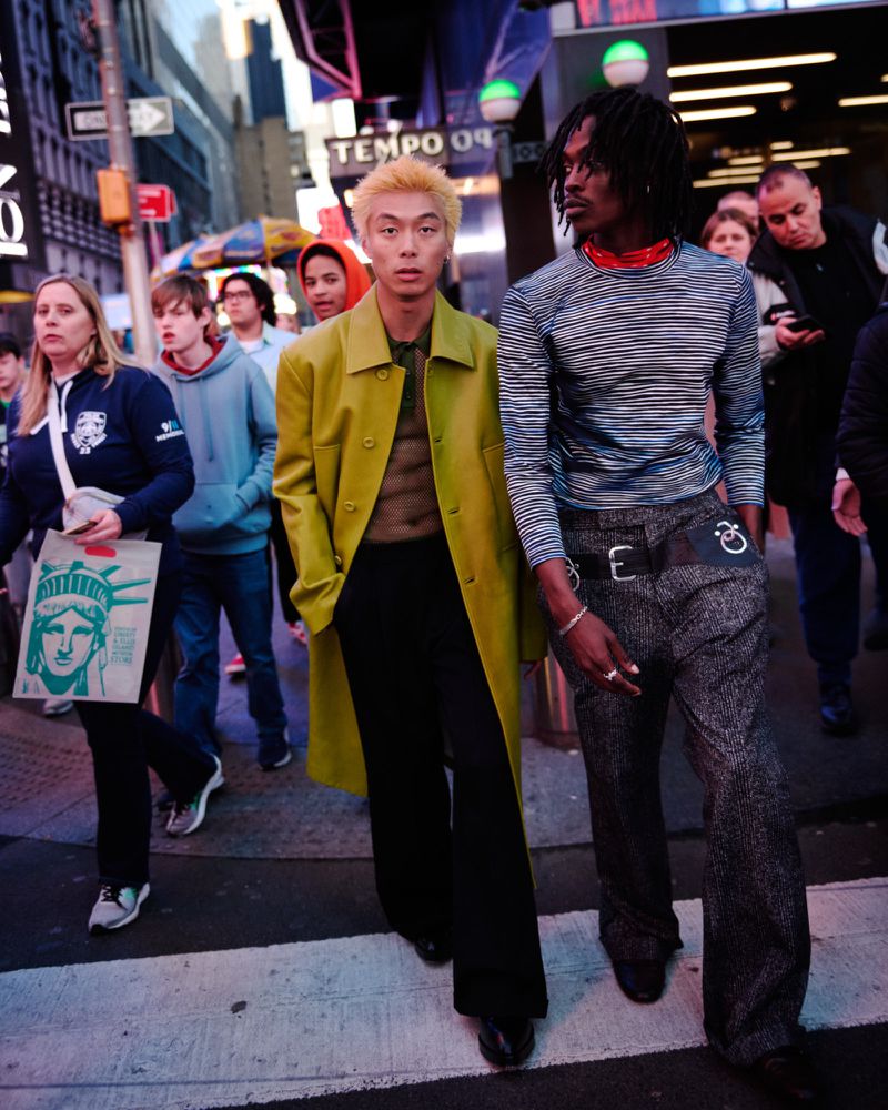 Two stylish adults cross a street in an urban setting, one with bright yellow coat and black pants, the other in striped shirt and patterned pants.