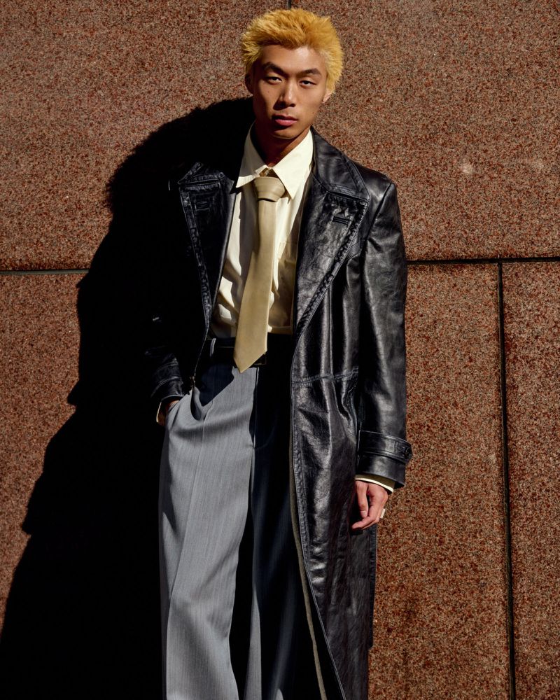 Young man with blond hair poses against a textured wall, wearing a black trench coat, shirt and tie, and gray trousers in natural light.