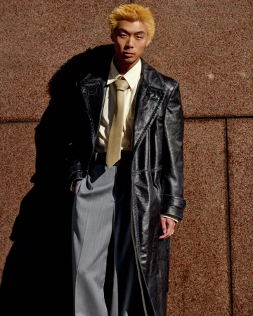 Young man with blond hair poses against a textured wall, wearing a black trench coat, shirt and tie, and gray trousers in natural light.