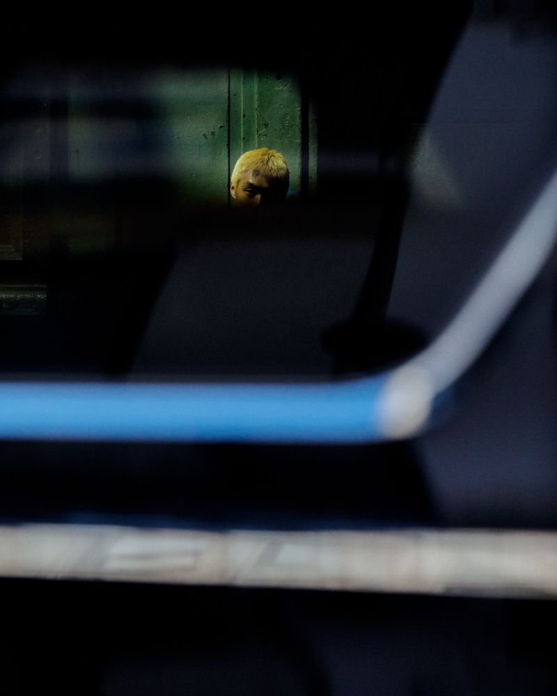 A person with dyed blonde hair framed through a dark window reflection with a blue accent bar.