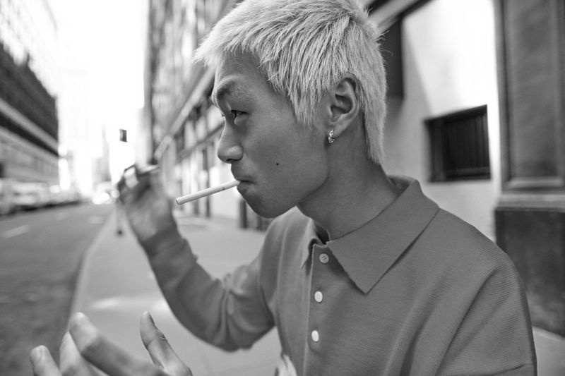 Black and white portrait of an Asian man smoking outdoors in a city, wearing a polo shirt, his light-colored hair contrasting with the monochrome.