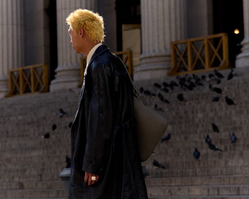 A man with blonde hair walks past a building facade with columns and stairs, with pigeons gathered on the steps.