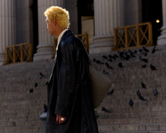 A man with blonde hair walks past a building facade with columns and stairs, with pigeons gathered on the steps.