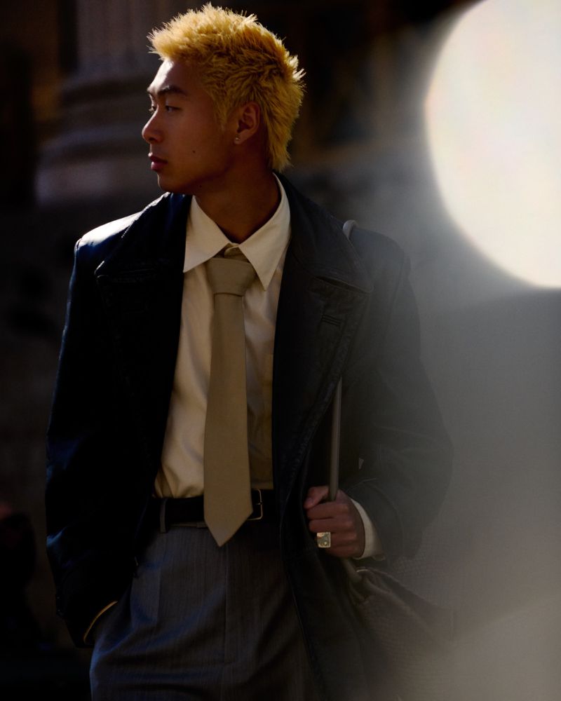 Stylish young adult in a dark coat, white shirt, and grey trousers. The fashionable man stands outdoors in sunlight, looking to the side.