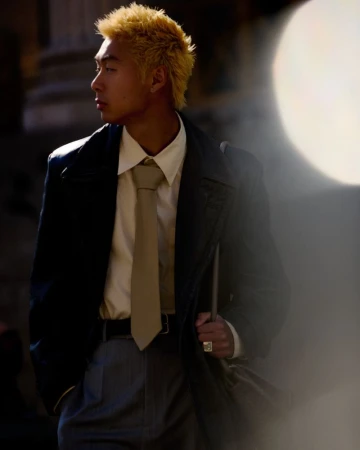 Stylish young adult in a dark coat, white shirt, and grey trousers. The fashionable man stands outdoors in sunlight, looking to the side.