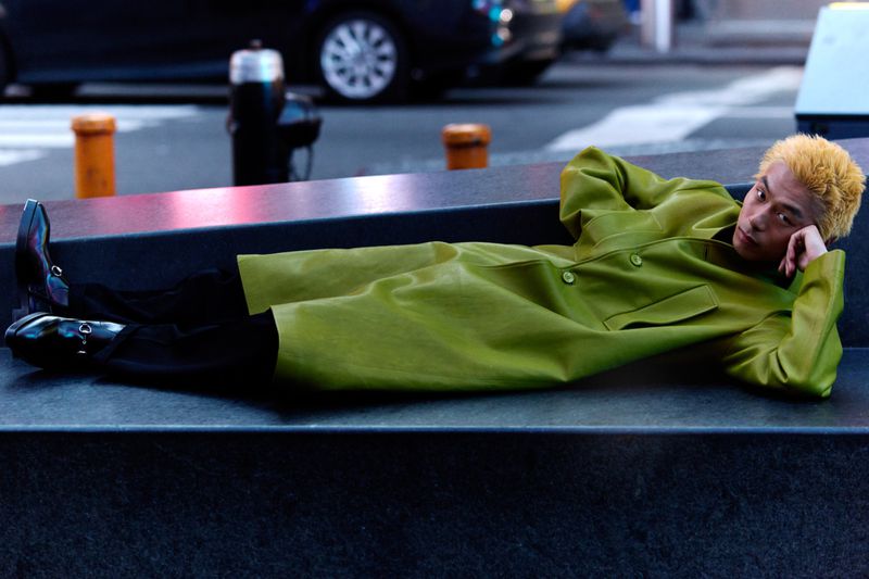 An Asian man with dyed blonde hair lies on a concrete bench in a vibrant green coat. Cars and urban features fill the background.