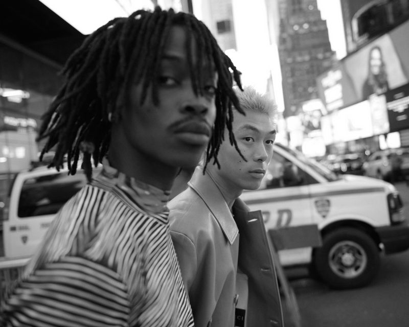 Black and white medium shot of two men in an urban setting with blurred city buildings in the background and a white van parked behind.