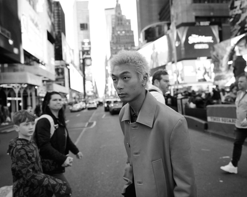Black and white photo of a young Asian man in a coat standing on a street in Times Square, New York City, surrounded by pedestrians.