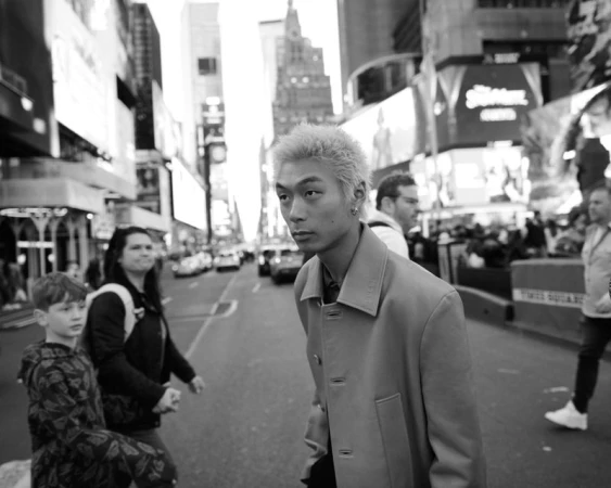 Black and white photo of a young Asian man in a coat standing on a street in Times Square, New York City, surrounded by pedestrians.