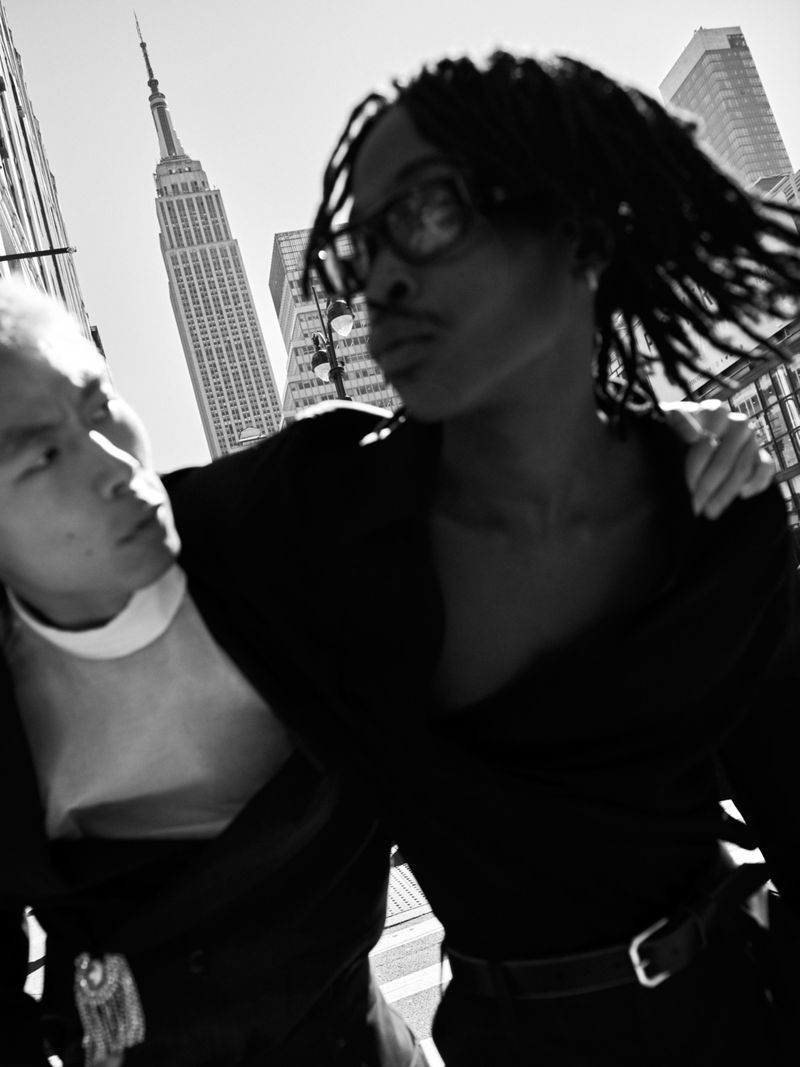 Black and white shot of two people in an urban setting, with a skyscraper in the background, creating a high-contrast scene.