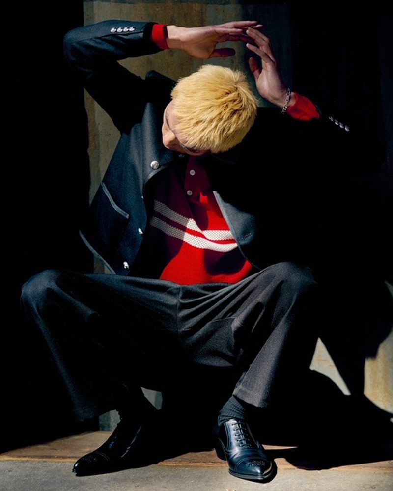 Stylish blond man in dark suit and black dress shoes strikes a pose against an industrial wall, casting sharp shadows. 