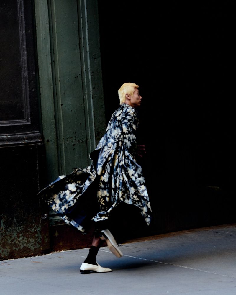 Model with blond hair and floral robe walks past a dark doorway on a city street in a fashion editorial style shot. 