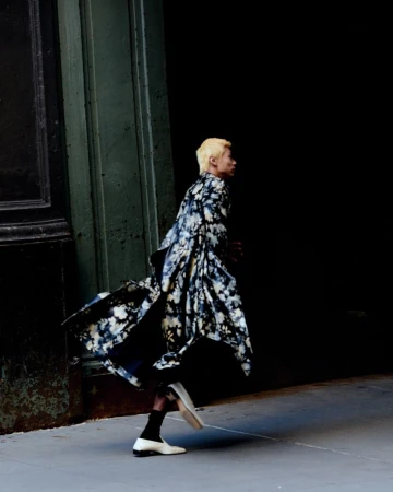 Model with blond hair and floral robe walks past a dark doorway on a city street in a fashion editorial style shot. 