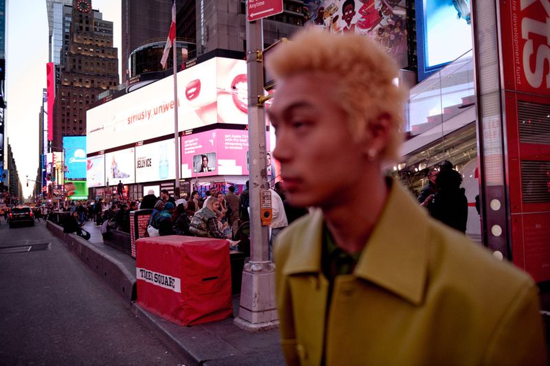 Candid medium shot of a blonde adult male in a yellow jacket against the backdrop of Times Square at dusk with illuminated billboards.