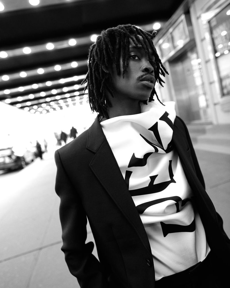 Black and white portrait of an adult male in a suit jacket and graphic hoodie standing outside in front of a building at night.