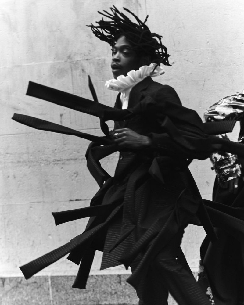 Black and white fashion photography of a Black male model wearing a ruffled collar and black coat with fabric strips.
