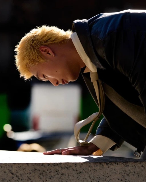 Close-up of an adult male with blonde hair wearing a dark jacket, standing outdoors with blurred greenery in the background. 