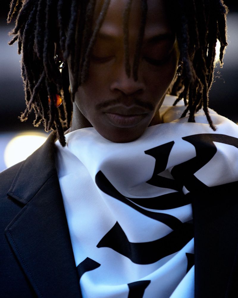 Portrait of a Black man with dreadlocks wearing a patterned white scarf and dark blazer
