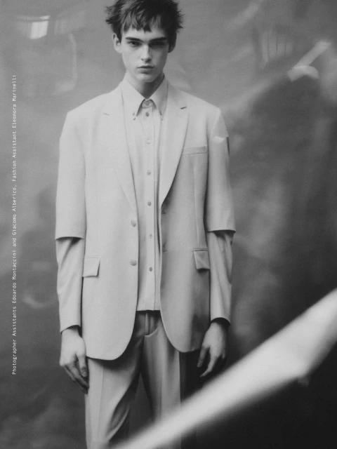 A young adult male model in a light-colored suit poses indoors against a neutral backdrop in a fashion editorial shot in black and white.