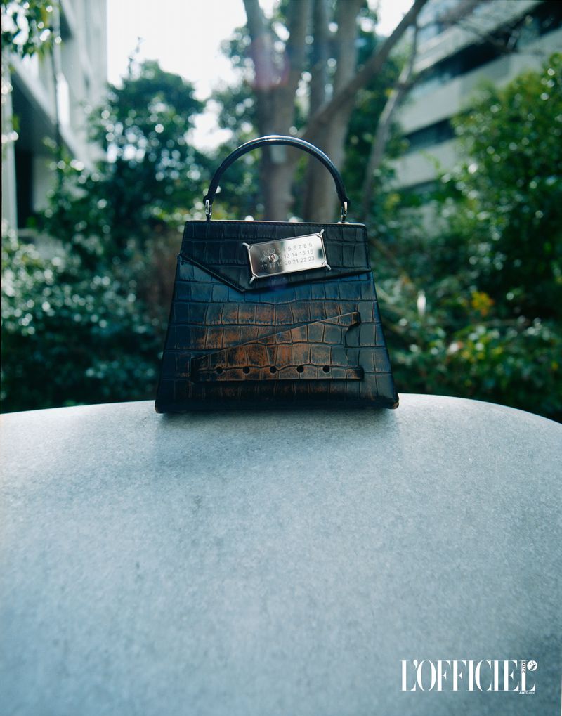 High-fashion crocodile handbag on a marble-like surface with soft, diffused outdoor lighting and a blurred cityscape background.