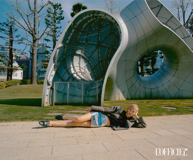Woman lies on pavement near modern metal sculpture in urban park on sunny day; she's wearing silver shorts and a gray jacket.