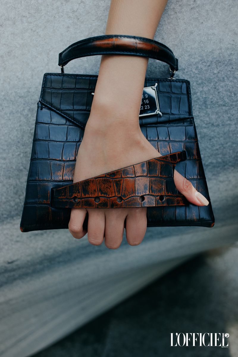 Fashion shot of a woman's hand holding a black crocodile-print leather handbag with a brown handle, against a grey backdrop.