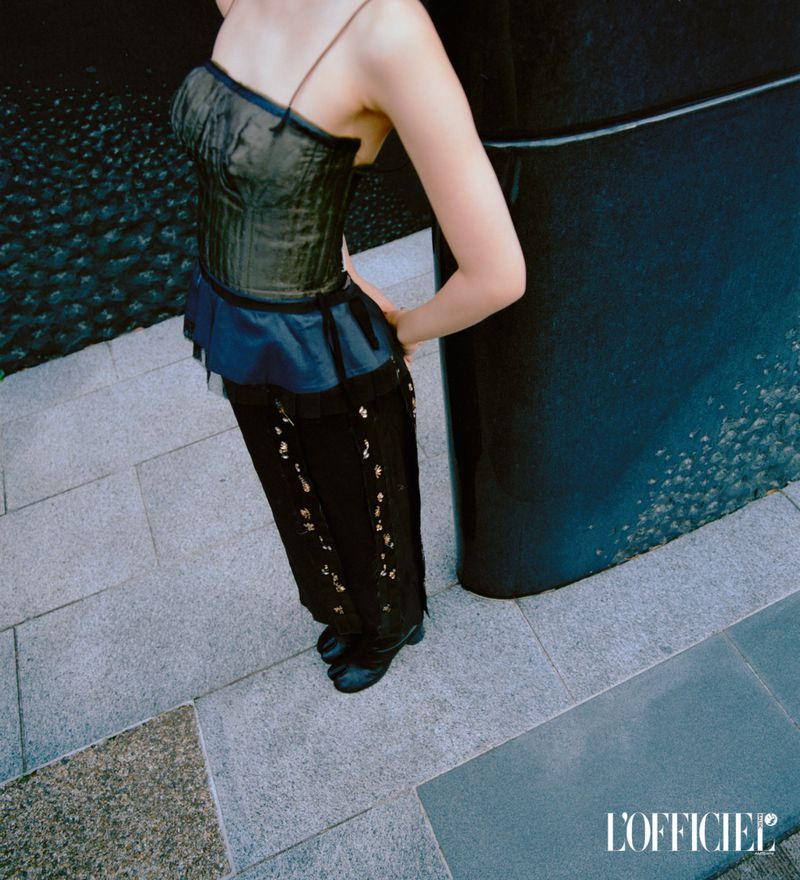 Woman wearing a corset top, pants with floral design, and Japanese-style shoes poses on a stone surface next to a dark pillar.
