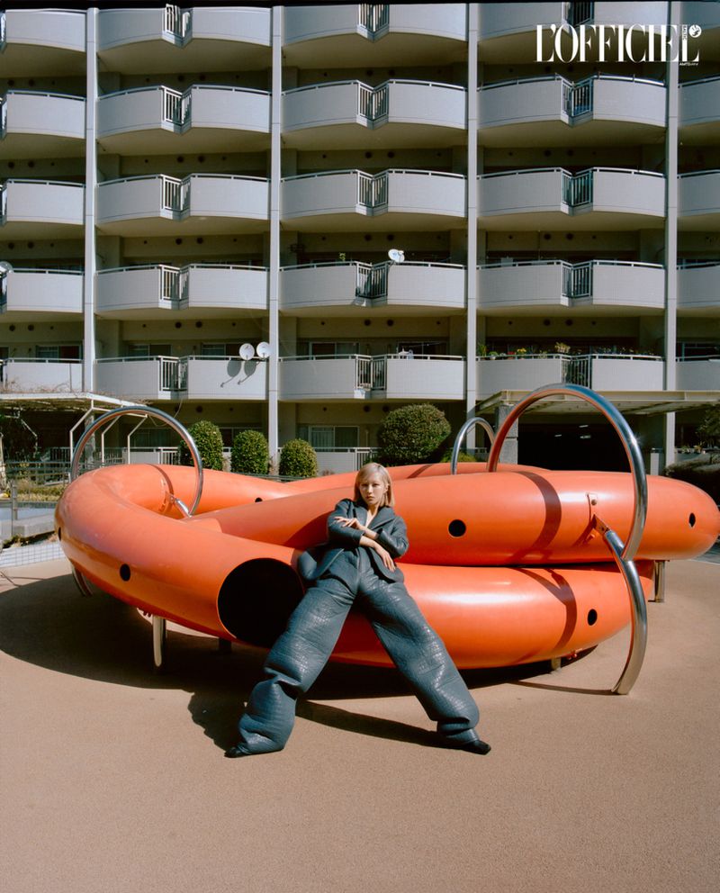 Fashion editorial featuring an adult female model posing near a large orange playground structure with an apartment building background.