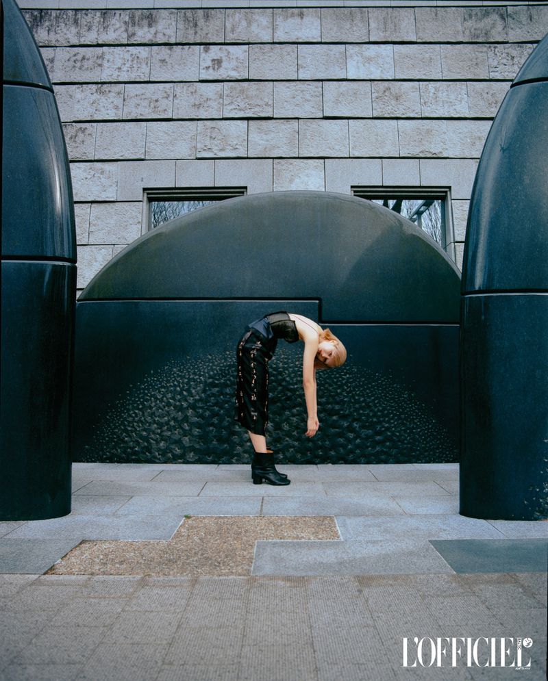 A woman in a black top and patterned culottes arches backward, her head down, in front of an abstract art piece with stone buildings.